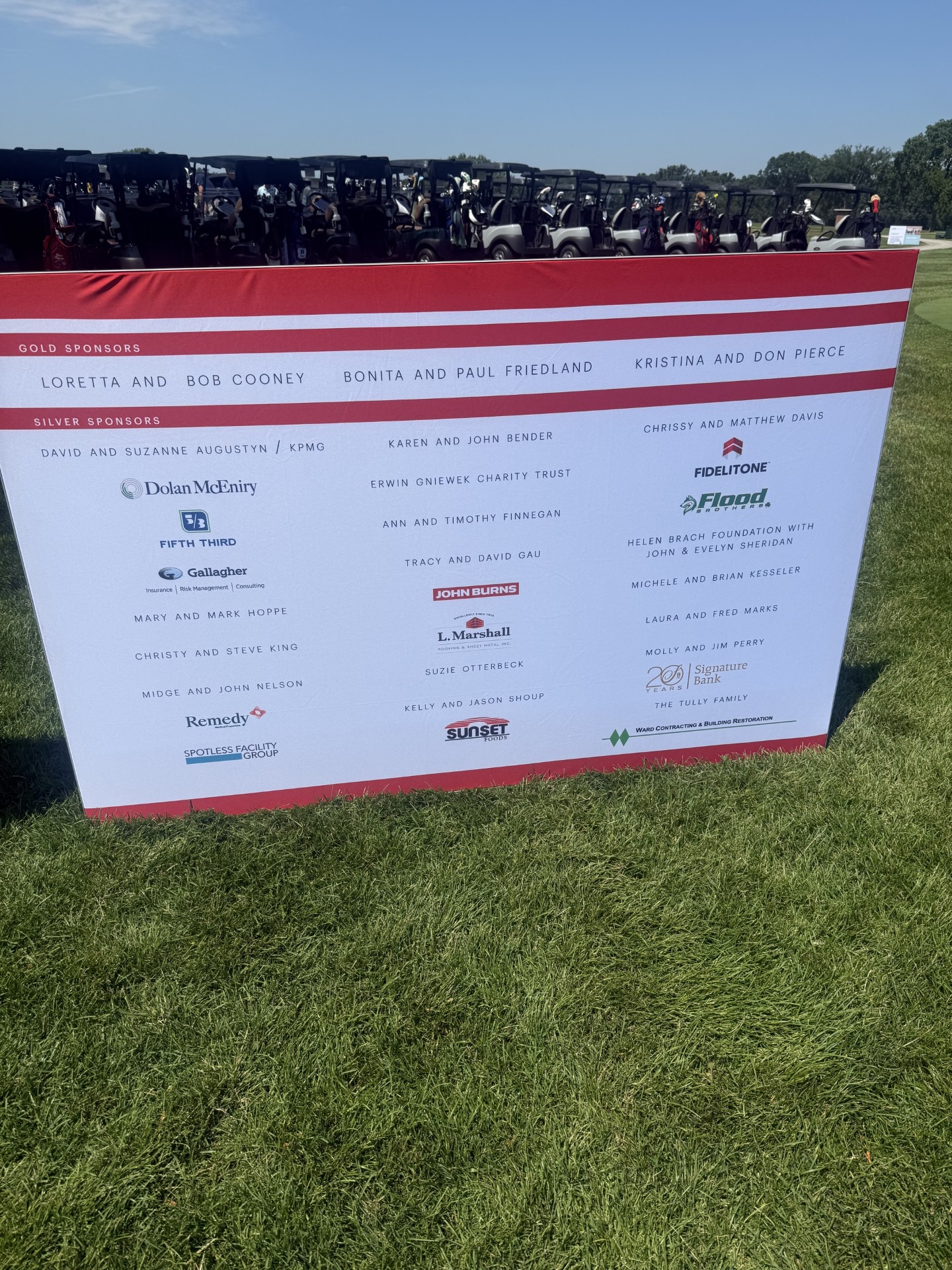 Gold and Silver sponsor board at the Catholic Charities Golf Classic, with golf carts lined up behind it