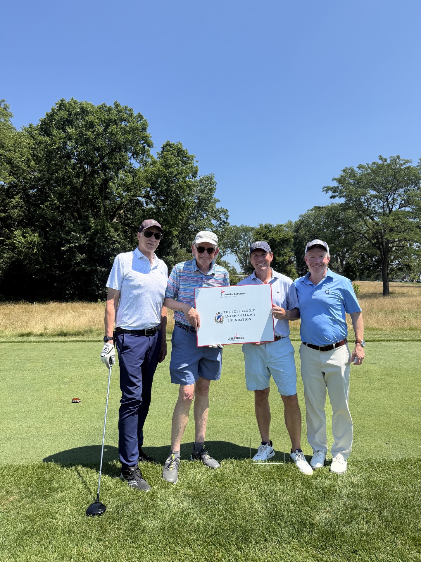 Four Pope Leo XIV American Legacy Foundation representatives hold the Foundation sign on the fairway at the Catholic Charities Golf Classic